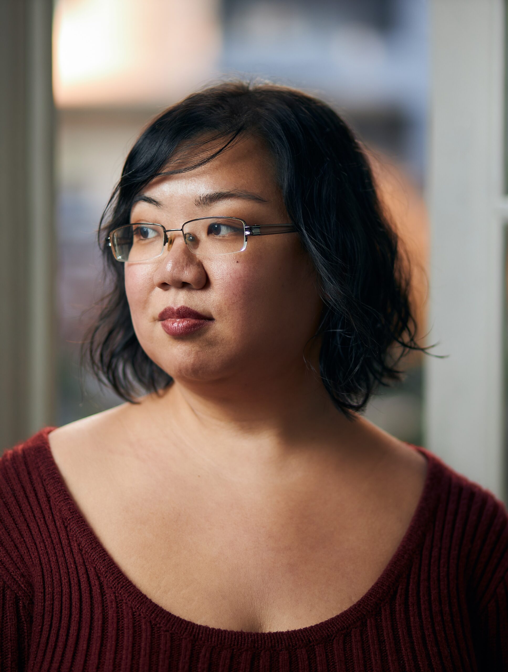 The image shows author Nghi Vo smiling softly while looking slightly to the side. She is standing in front of a blurred natural background with greenery. Nghi is wearing a patterned shawl over a dark top, and her long dark hair is draped over one shoulder.