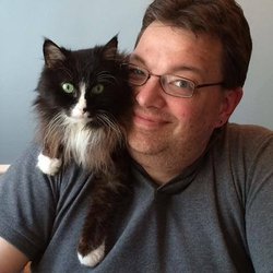 Jim Johnson is pictured in front of a neutral gray background, smiling slightly with a calm, direct expression. He wears black rectangular glasses, a dark collared shirt, and a charcoal jacket. A cat rests on his shoulder, with a black face and legs, tan and white chest, and white paws, adding a striking contrast to the darker clothing.