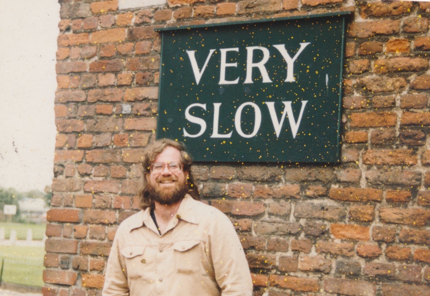 Greg Ketter is standing outdoors in front of a stone wall with a green sign that reads "Very Slow." He has shoulder-length brown hair and wears a light tan button-up shirt. His expression is relaxed and slightly smiling, giving a friendly, approachable impression.